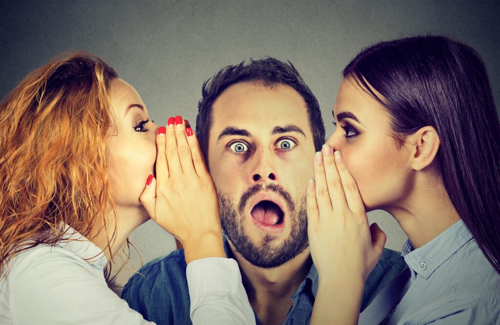 Two women whispering secret gossip in the ear of an amazed, shocked man.